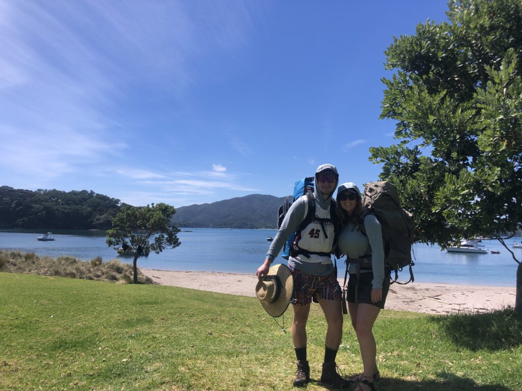 A couple stands in the grass in front of a sandy beach. They beach has sparkling blue water and there is blue skies above, they are camping on Urupukapuka Island in New Zealand