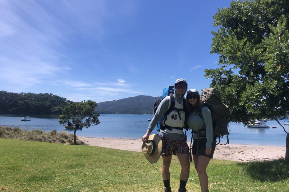 A couple stands in the grass in front of a sandy beach. They beach has sparkling blue water and there is blue skies above, they are camping on Urupukapuka Island in New Zealand