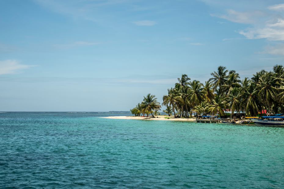 San Blas Island sailing trip. The island has crystal clear Caribbean blue water with blue skies overhead. Photo shows an island on the righthand side covered in thick tropical trees