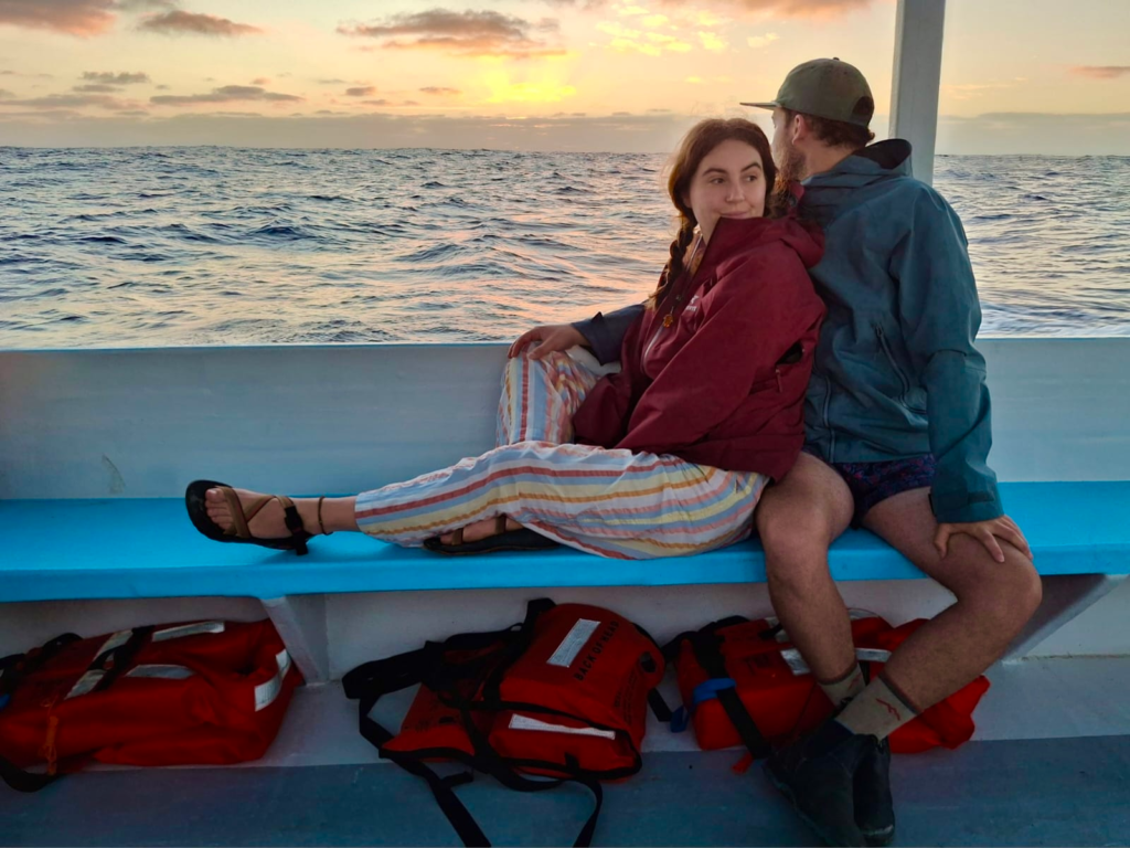 A couple sits on a bench on a boat, the sun is rising over the water behind them. The man is looking at the sunrise and the woman is looking off in the distance behind the camera