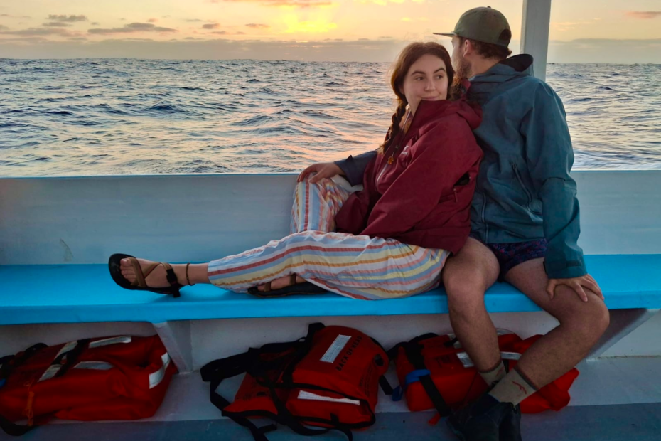 10 essential items for traveling to tonga. A couple sits on a bench on a boat, the sun is rising over the water behind them. The man is looking at the sunrise and the woman is looking off in the distance behind the camera