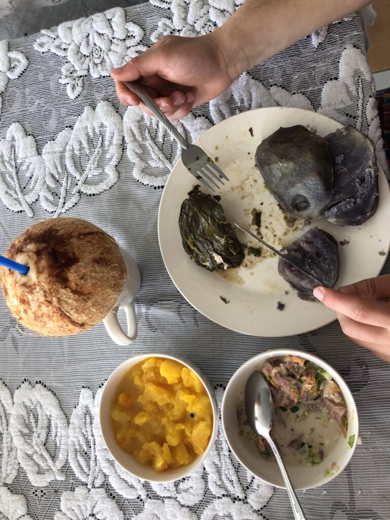 An overhead view of a traditional Tongan meal on a lacy table cloth. A coconut with a straw in it, a bowl of cut papaya and a plate of cut being cut up