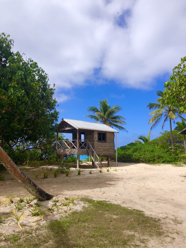 2 week tonga travel itinerary, a resort in Tonga. Sandy beach with a fale standing on stilts. Palm trees frame the sides and there are blue skies above