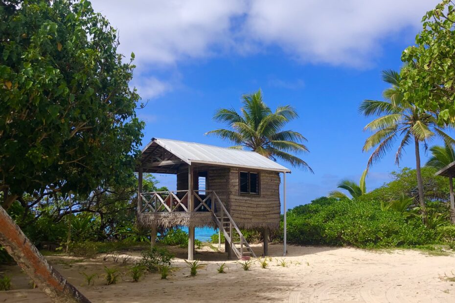 2 week tonga travel itinerary, a resort in Tonga. Sandy beach with a fale standing on stilts. Palm trees frame the sides and there are blue skies above
