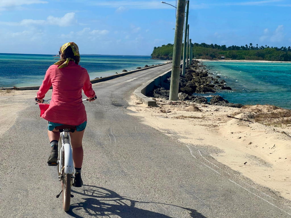 Resort in Tonga activity. A woman biking along a low bridge that has beautiful blue water on either side and an island across the way. She's wearing shorts and a pink longsleeve top