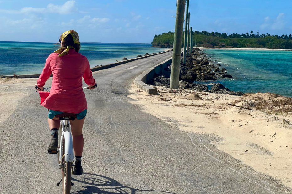 Resort in Tonga activity. A woman biking along a low bridge that has beautiful blue water on either side and an island across the way. She's wearing shorts and a pink longsleeve top. tonga budget 2025