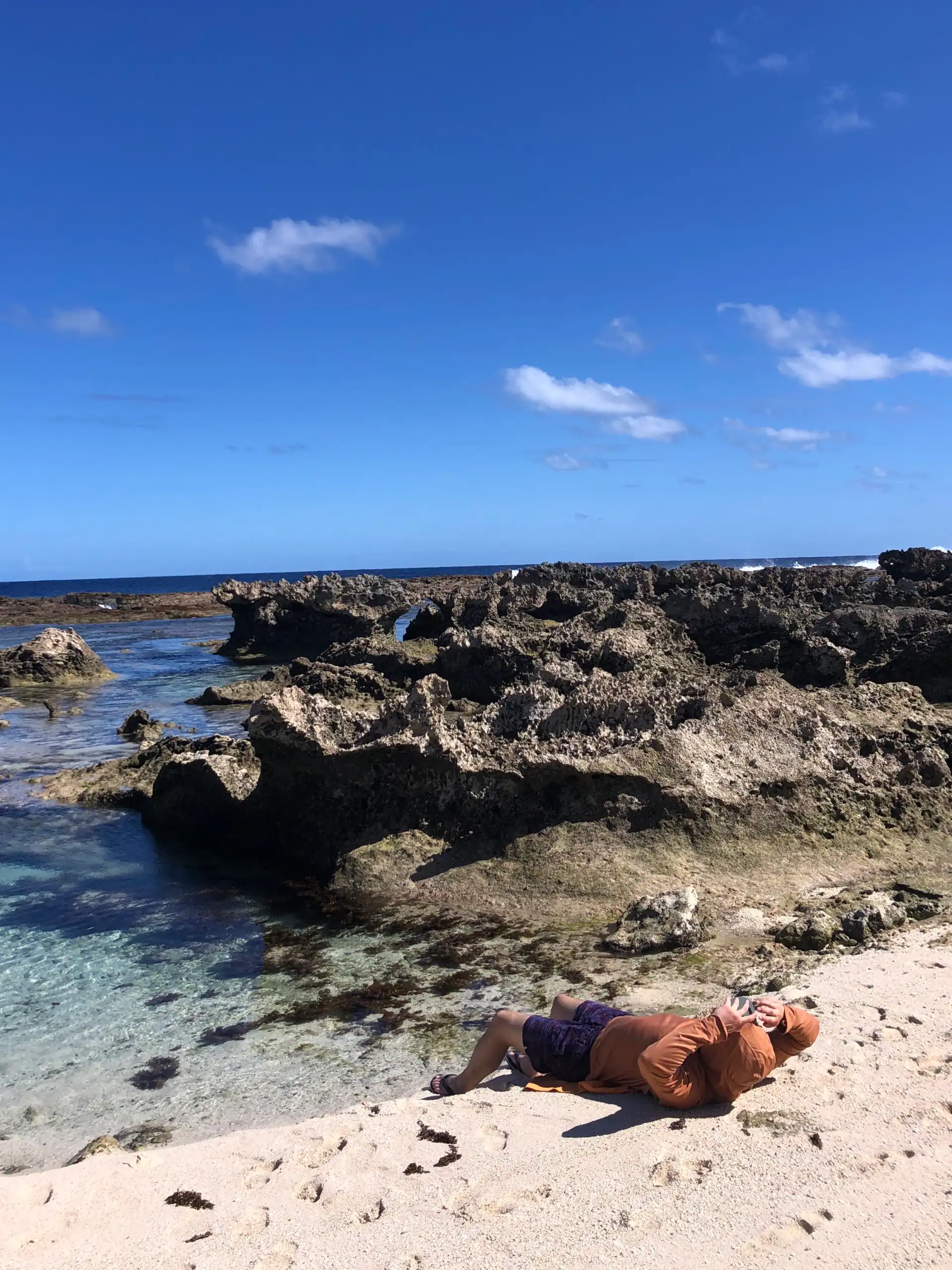 Planning a trip to Tonga. Man relaxing on sandy beach on 'Eua with rocky volcanic flows in front of him