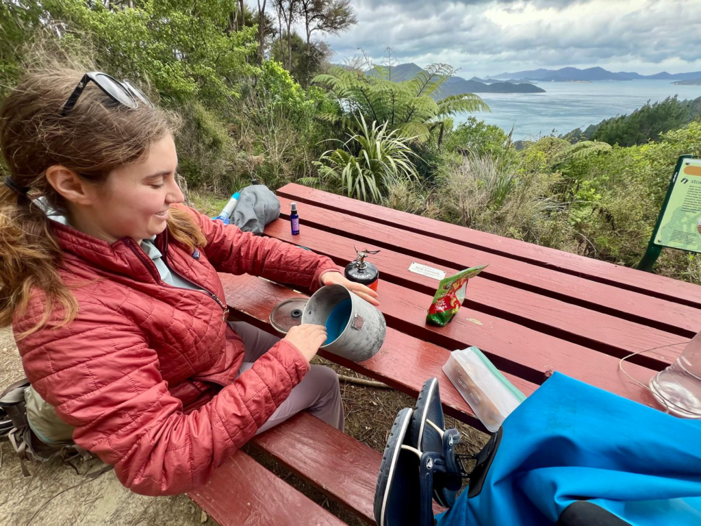 woman at a table on the queen charlotte track cooking a meal with a backpacking stove