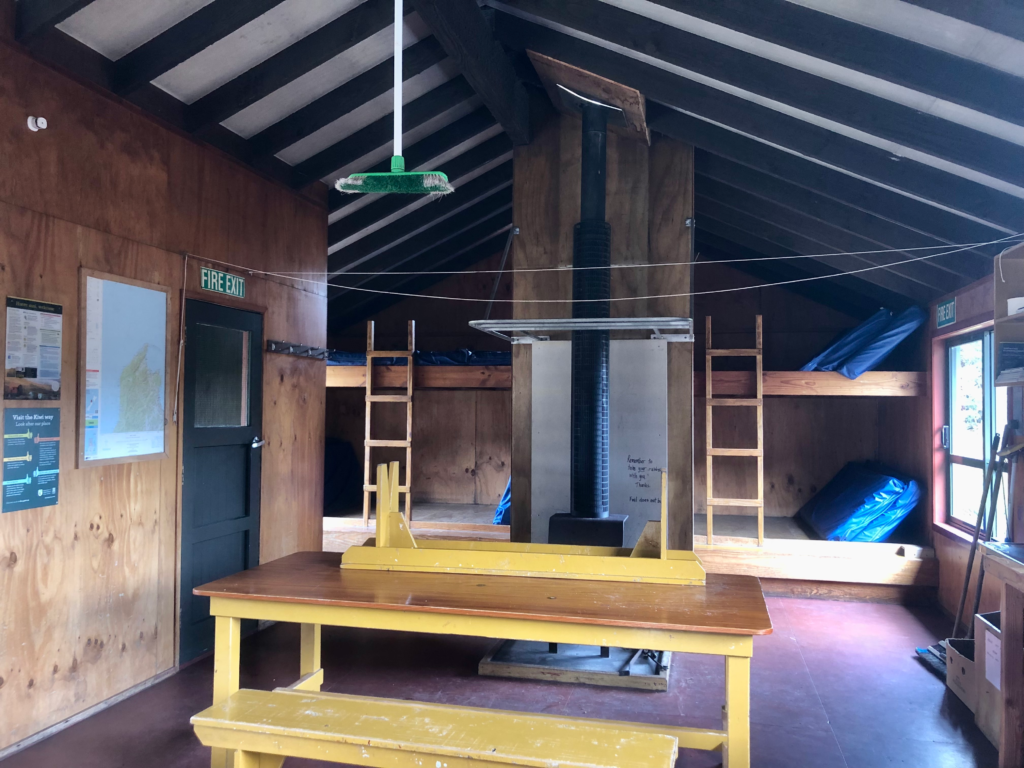 inside saxton hut on the heaphy track. wooden table in middle of room with fireplace and bunks behind it