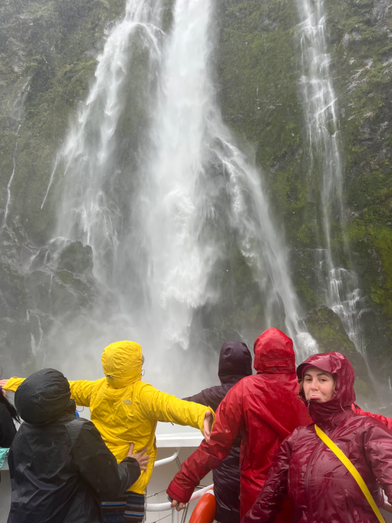 1 week south island road trip isn't compelete without a trip to milford sound. 5 women in rain jackets in front of a massive waterfall