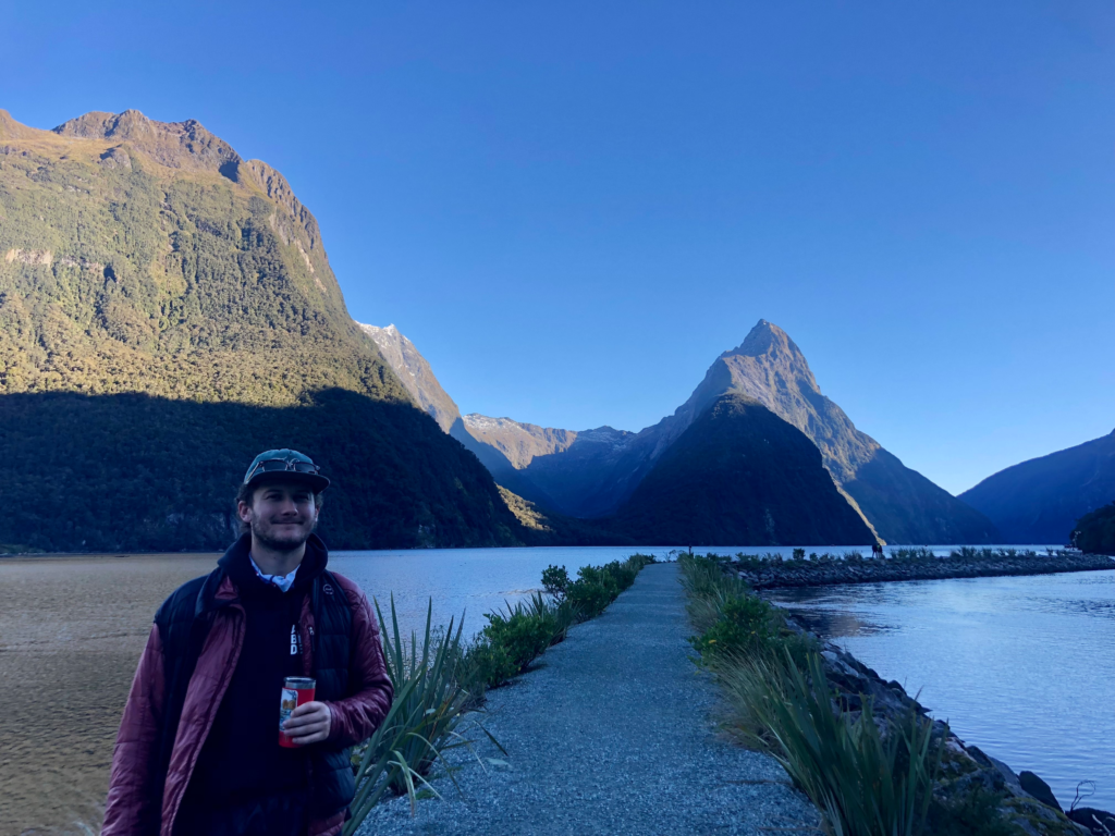 man standing in front of water with mitre peak mountain in background in milford sound new zealand