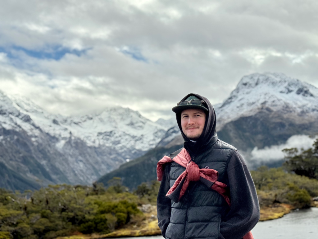 how to spend 1 week onthe south of island of NZ, go to the milford sound. man standing in the snow capped mountains smiling on a walking trail