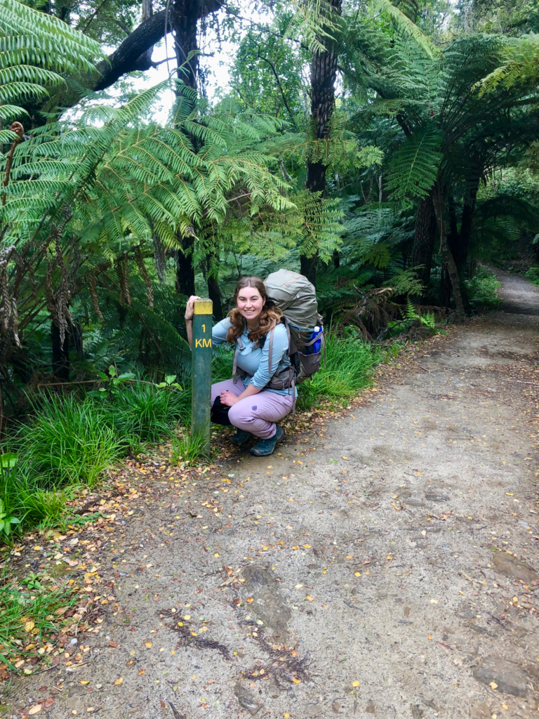 ultimate guide to hiking the queen charlotte track in 4 days, woman crouching next to sign post that says 1 km. She has a big backpack on