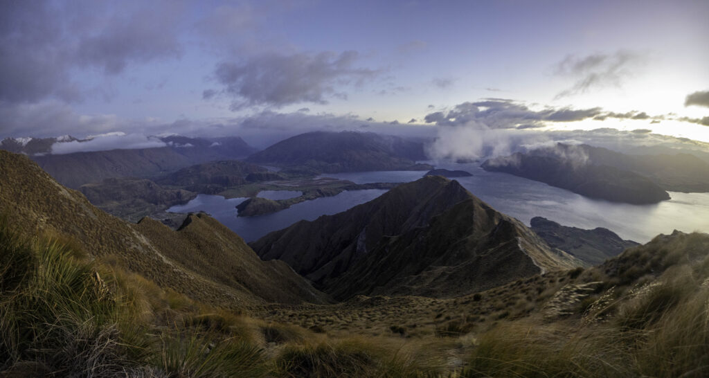 1 week south island new zealand road trip itinerary, 2 days in wanaka. view from roys peak of shrub covered mountains down to a blue lake