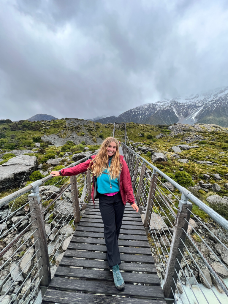1 week south island new zealand road trip itinerary, stop for a hike in mt. cook. woman standing on bridge on cloudy day, alpine shrubbery in background
