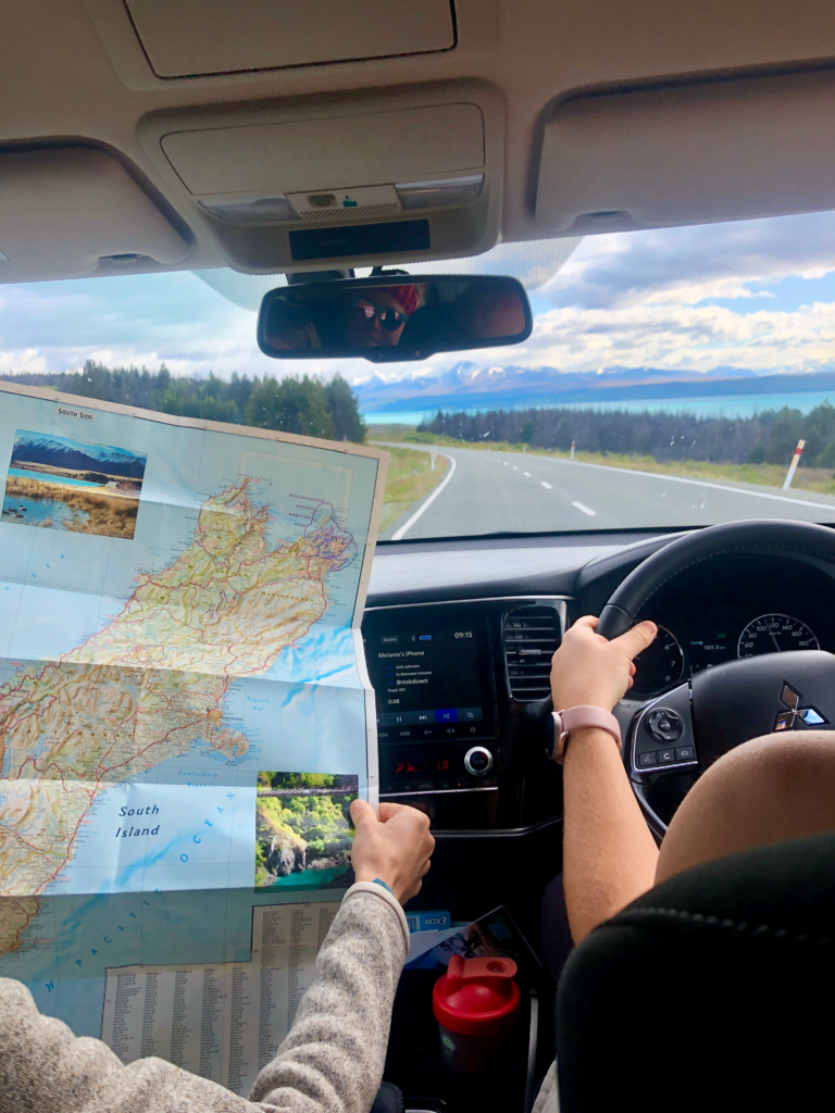 1 week south island road trip itinerary. view out car windshied of a map of new zealand while someone drive down a country road