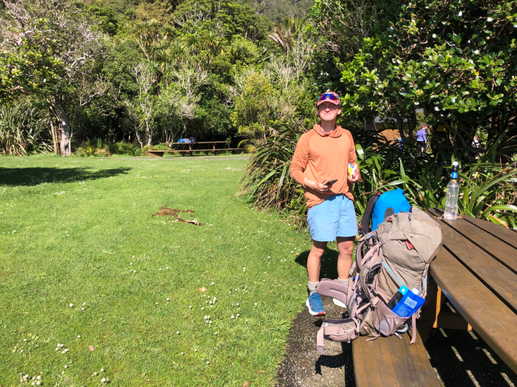 guide to hiking the queen charlotte track in 4 days man in hiking clothes standing on green grass next to picnic table and backpacking backpacks