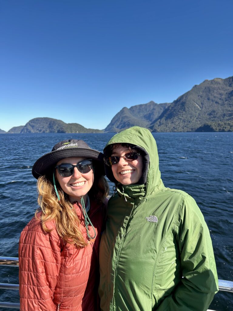 2 women in doubtful sound new zealand, smiling in rain gear in the sun, ocean in background