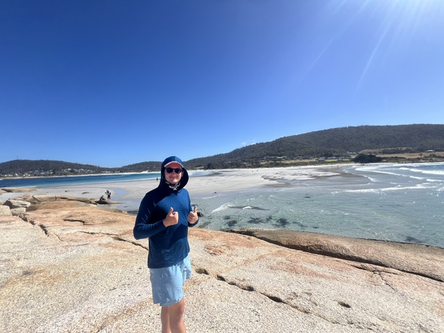 top 8 reasons to visit tasmania - amazing beaches. man standing on white and red rocks in the bay of fire tasmania, crystal clear water and white sand beach in background