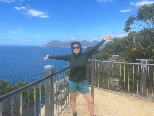 top 8 reasons to visit tasmania! woman standing with arms out wide at cape tourville lighthouse on a sunny day with blue skies